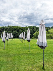 End of the Season: Rows of Closed Beach Umbrellas on a Green Lawn Under a Cloudy Sky. Awaiting the Sun.Off-Season Scene: An Empty Recreation Area with Closed Umbrellas on a Cloudy Day
