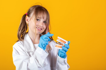 Dental health and teethcare concept. Young girl in dentist uniform and rubber gloves holding human jaw model with dental mirror on yellow background. Can be used to represent Dental clinic for kids