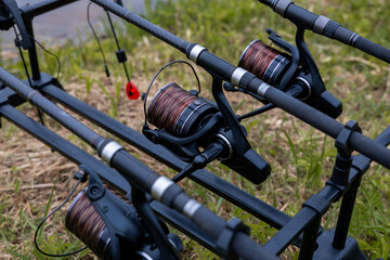 Close up of fishing rods mounted on a pod with reels and brown line, positioned on grassy terrain near water, featuring a red bite alarm. © True Pixel Art