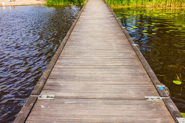 Wooden floating pier with metal hinges crossing over calm lake water with green reeds and lily...