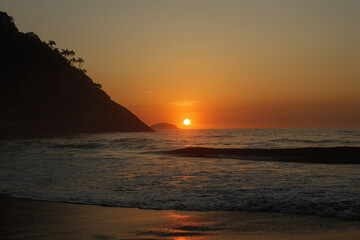 Nascer do sol na praia do Leme, Rio de Janeiro. Céu alaranjado e mar calmo, com uma montanha com silhuetas de coqueiros à direita projetando-se em direção ao mar e um pequeno morro ao fundo. © SONIA