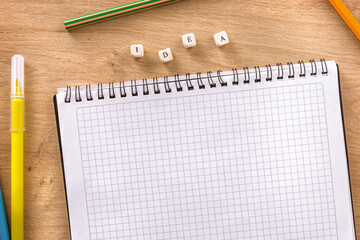 Top view of empty squared notebook with colorful pencils and wooden cubes forming the word idea on desk background