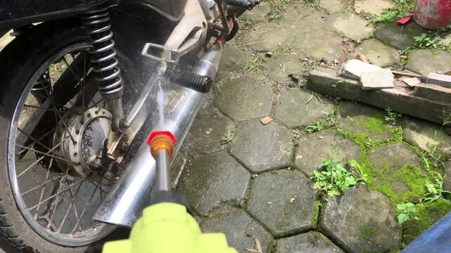 A person washes a motorcycle using a high-pressure spray gun. Water blasts away dirt and grime, making the bike clean and shiny in this routine cleaning activity.