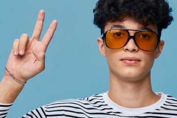 young man wearing orange sunglasses and striped shirt shows peace sign with fingers against blue...