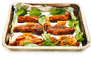 Raw uncooked pork belly portions, tangerine segments and basil fresh herb on cooking foil and metal cooking tray, white background. Marinated meat ready to be cooked in an oven.
