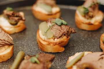 Gourmet beef canapes garnished with pickles and herbs on a black serving plate, perfect for events and catering.