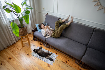 Bored woman with feet up on couch absorbed in smartphone, browsing internet chats and social feeds while cat lies nearby. Laziness, digital dependency, emotional detachment, indoor inertia, solitary
