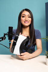 Female tech blogger seated at desk, reviewing a VR headset on camera. She speaks into a microphone...