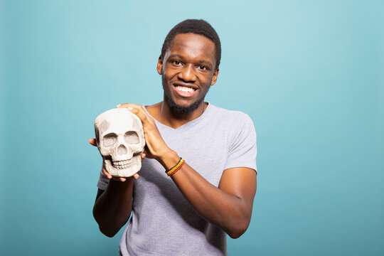 Smiling man holding and presenting a human skeleton head towards the camera in a blue background studio. Young black guy happily grasping a skull and standing against an isolated background.