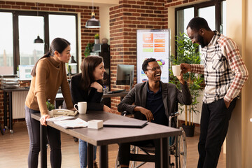 Group of diverse coworkers, including a wheelchair using business analyst, happily bonding in an office environment. They share stories and laughter during their break at a startup company.