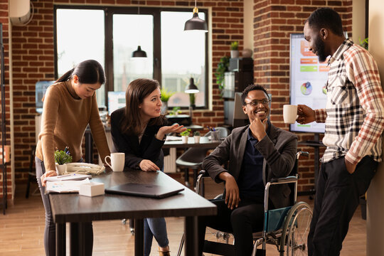 African american man in a wheelchair enjoys a lighthearted conversation with his diverse coworkers during a break. Multiethnic colleagues laugh together in a brick wall office, sharing stories.