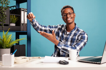 African american male consultant seated at desk, gesturing towards set of keys on his hand. Young smiling man looking at camera and showing his office key, representing new business achievement.