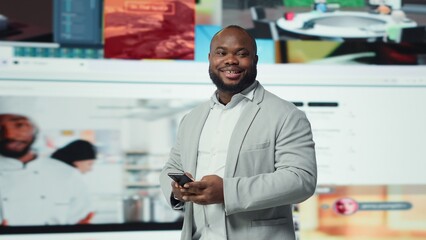 Portrait of smiling man browsing social networks feed on smartphone in front of videowall. Jolly african american person uses phone, floating icons highlighting interactive engagement online, camera A
