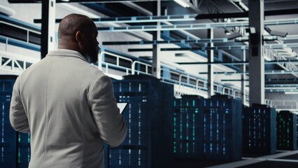 Data center system administrator checking infrastructure diagnostics using tablet screen. IT specialist walking in high tech server room, examining data charts during system audit, camera A