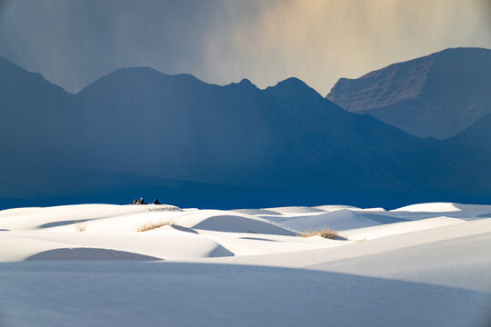 White rolling sand dunes and dark mountains in the distance under a rain cloud at White Sands National Park, New Mexico. A group of hikers can be seen in the distance.