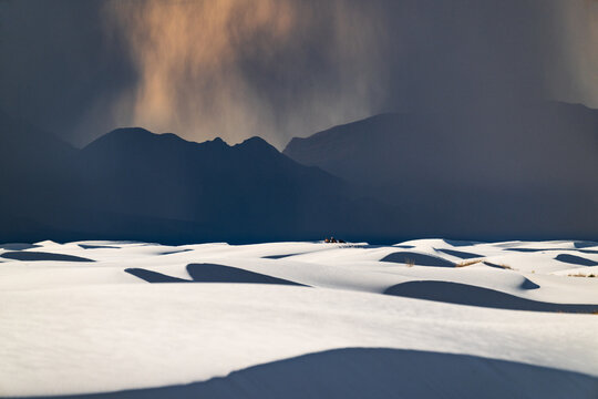 White rolling sand dunes and dark mountains in the distance under a rain cloud at White Sands National Park, New Mexico. A group of hikers can be seen in the distance.