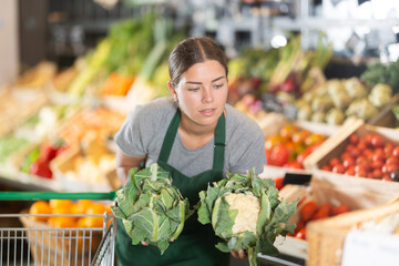 Sales girl works in vegetable store bio supermarket and goes through goods. She holds cauliflower...
