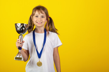 Happy smiling young blonde girl with a gold medal in white T-shirt holding winner cup or trophy on yellow background. Champions, competition and sports concept