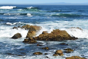 Powerful ocean waves crash dramatically against rugged coastal rocks under a bright blue sky. 