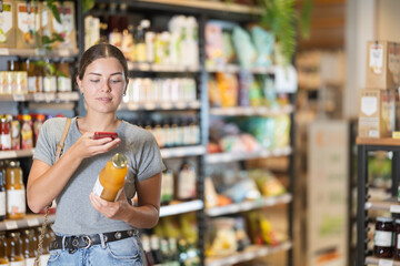 Interested young girl scanning QR code on juice bottle to access detailed product information while shopping in organic grocery store. Concept of using modern technology to help buyer..