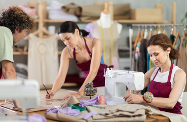 Friendly tailor and tailoress drawing on paper and speaking as other working with sewing-machine