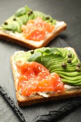 Delicious sandwiches with avocado, salmon, cream cheese, basil and capers on black table, closeup