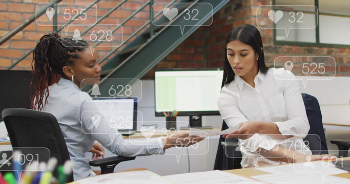 Handing document, businesswomen wearing suits collaborating at office desk with social media icons - Powered by Adobe