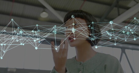 Speaking adult Asian woman holding smartphone near mouth in hangar, with digital network overlay