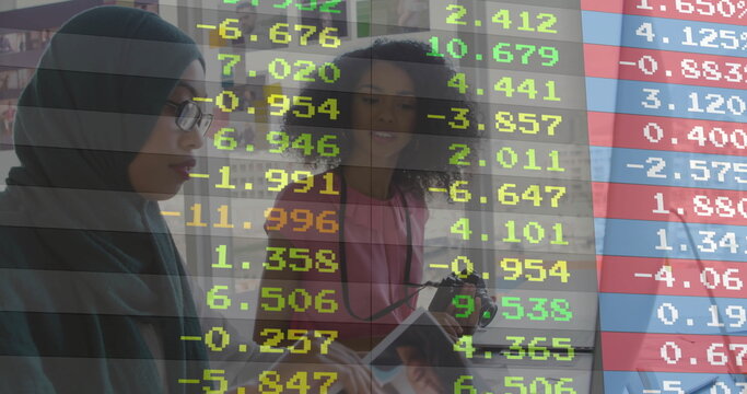Reviewing financial data overlay on tablet, analysts in hijab and pink top working in meeting room - Powered by Adobe