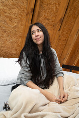 A smiling young woman sits on a cozy bed, wrapped in a blanket, enjoying a peaceful morning in a triangular glamping tent filled with soft light and rustic charm.

