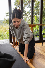 A young woman kneels beside a wooden bed in a cozy cabin with large glass windows. The scene evokes calmness, order, and a deep connection with nature.
