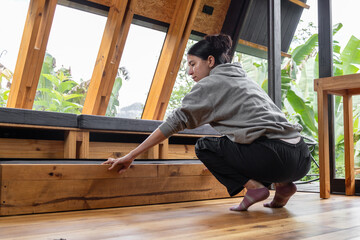 Seen from behind, a young woman squats as she opens a drawer beneath a wooden bed in a cozy cabin. The scene captures a quiet domestic moment, emphasizing order, presence, and harmony with the natural