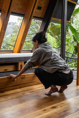 A young woman is seen from behind as she squats inside a rustic wooden cabin. Her posture suggests focus and calm, blending with the warm and peaceful atmosphere of this quiet mountain retreat.

