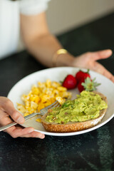 woman's hands holding healthy breakfast plate with avocado toast scrambled eggs and strawberries on her kitchen counter
