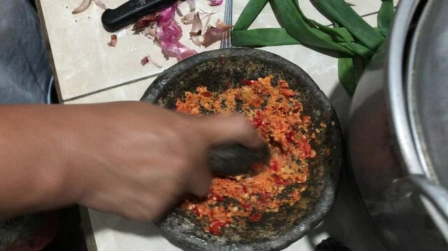 A woman is grinding spices using a traditional stone mortar and pestle, known locally as cobek and ulekan, a common cooking method in Indonesian households.