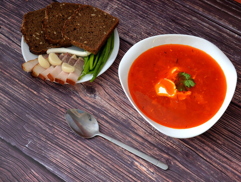 Borscht, tasty and nutritious soup, next to it a plate with rye bread, smoked lard, garlic and green onions and a tablespoon on a wooden table.