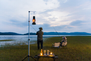 Romantic couple fishing together by the lake at dusk, sitting on camping chairs with lanterns and...