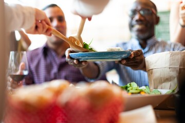 Group dining, sharing food. Diverse people enjoying a meal together. Hands serving salad. Community, togetherness, and food sharing at a table. Happy friends eating lunch, serving each other food.