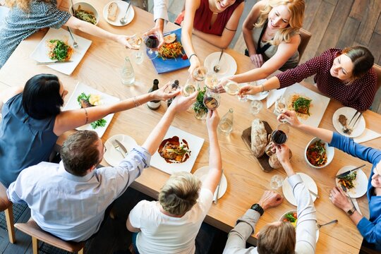 Diverse group of people enjoying a meal, raising glasses in a toast around a wooden table. Celebrating with food, drinks, and laughter. Diverse people toasting champagne over the table to celebrate