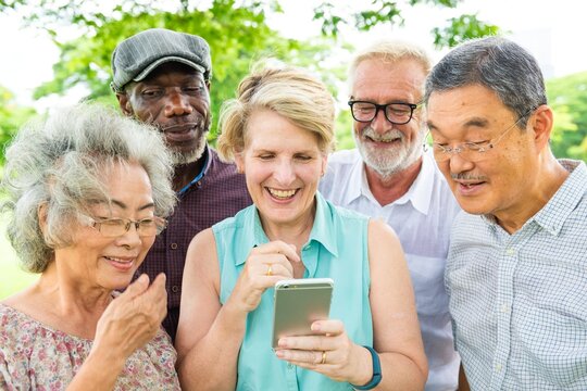 Group of diverse elderly people smiling at a smartphone. Elderly friends enjoying technology. Happy elderly group bonding over a phone outdoors. Diverse senior people using social media.