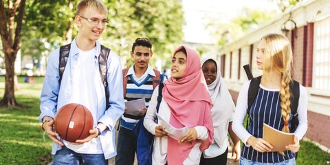 Diverse group of students walking outdoors, holding books and a basketball. Students include young men and Muslim women engaged in conversation. Diverse students walking outdoor