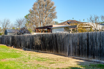 A worn and leaning timber boundary fence separates a grassy public green space from a row of...