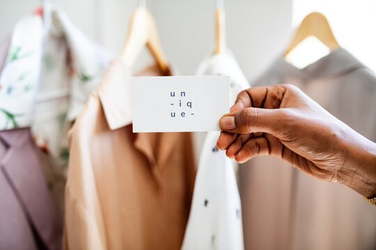 Close-up of a hand holding a card with 'unique' text, in front of hanging clothes. Fashion, style, and unique design elements are highlighted. Fashion business. Small business entrepreneur.