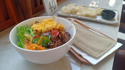 A delicious plate of black pepper barbecue beef served over steamed rice, accompanied by scrambled eggs and a side of fresh vegetable salad. A hearty and flavorful meal.