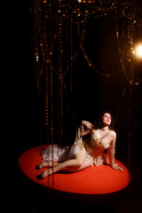 Young Caucasian woman in ornate golden corset, lying on red round stage floor, under hanging beads,...