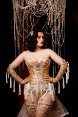 Young Caucasian woman in ornate cabaret outfit with golden corset, standing on dark stage under chandelier, front view. Concept: cabaret, glamour, theatrical show, vintage performance.