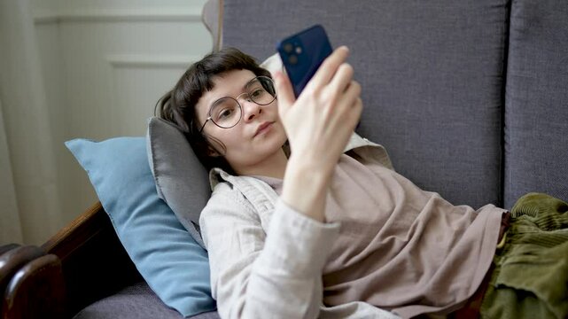 Relaxed woman browsing social media on smartphone while lounging on sofa, absorbed in digital web feed. Device dependency, endless scrolling habit, internet downtime, modern leisure, lonely isolation