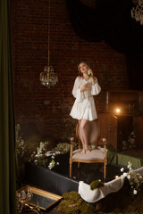 Young Caucasian woman in romantic white dress sits barefoot on chair in surreal studio swamp, eyes closed, front view. Moss, water, brick wall. Concepts: introspection, creativity, fine art, dream.