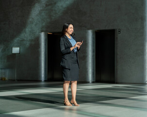 Obraz premium Smiling Asian businesswoman using smartphone in modern office lobby. Confident young professional walking with phone in elegant corporate interior