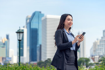 Successful Asian businesswoman using phone outdoors with skyscrapers in background. Young professional woman smiling in modern city district.